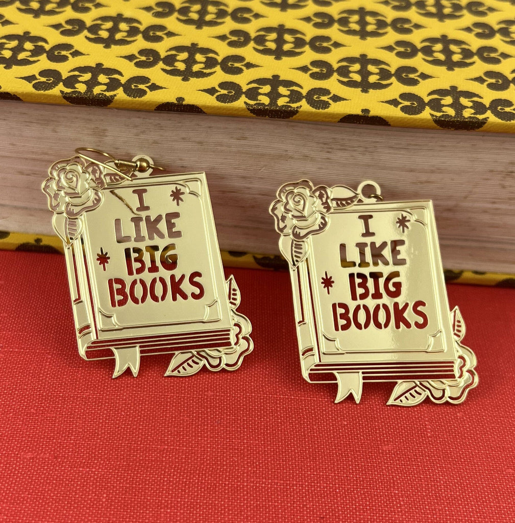 A pair of dangle brass earrings displayed on a book. The earrings read I Like Big Books.