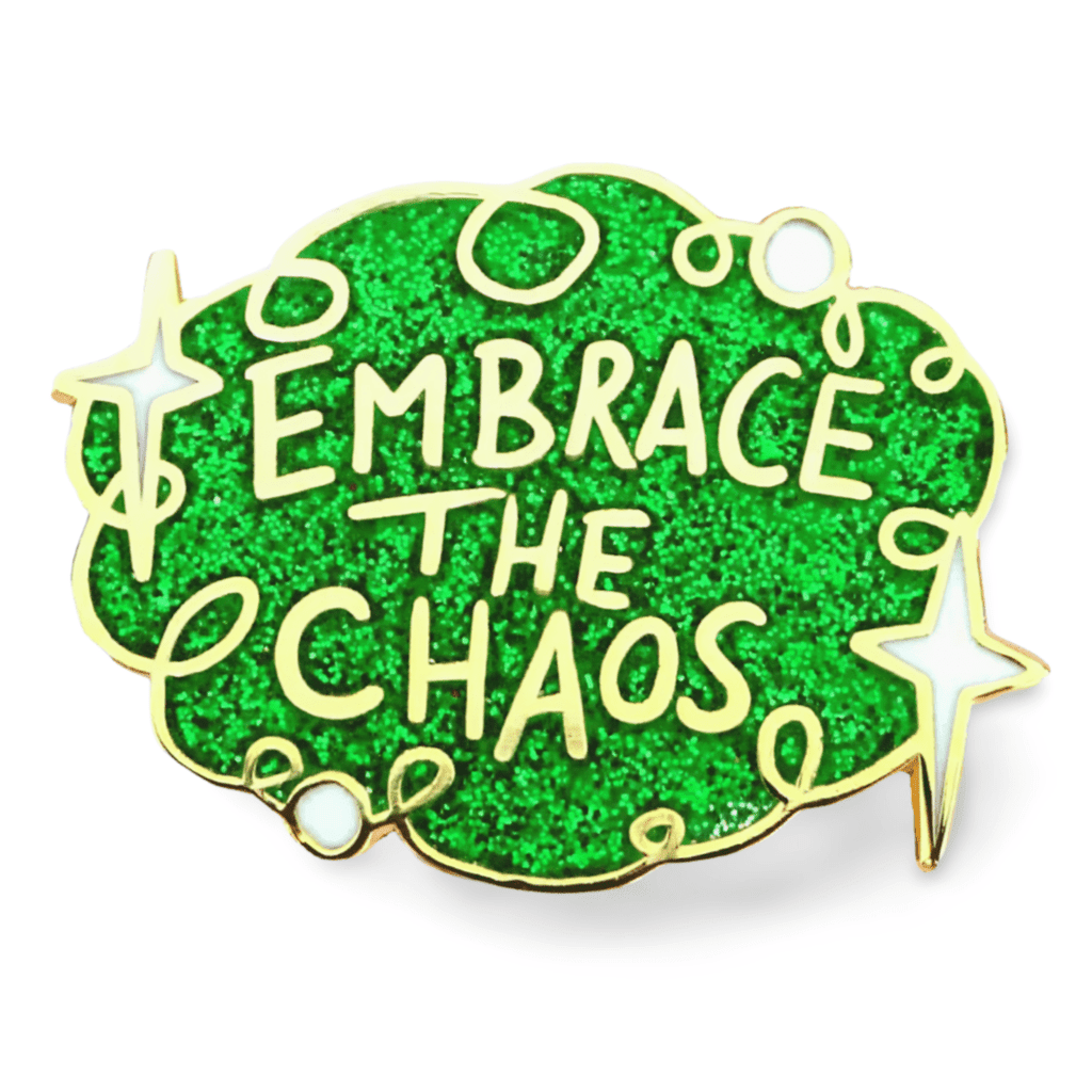 A hard enamel lapel pin on a white background. The pin is green glitter with white stars and reads Embrace the Chaos.