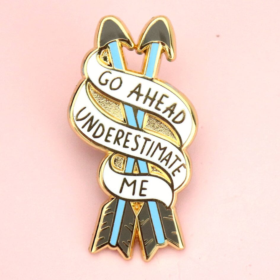 A hard enamel lapel pin on a pink background. The pin is in the shape of two arrows and reads Go Ahead Underestimate Me.