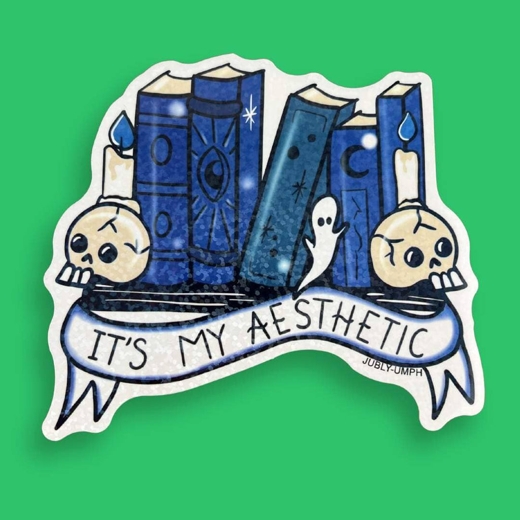 The vinyl sticker is in the shape of books on a shelf with skulls and candles. It is on a green background. The sticker is blue with glitter and reads It's My Aesthetic.