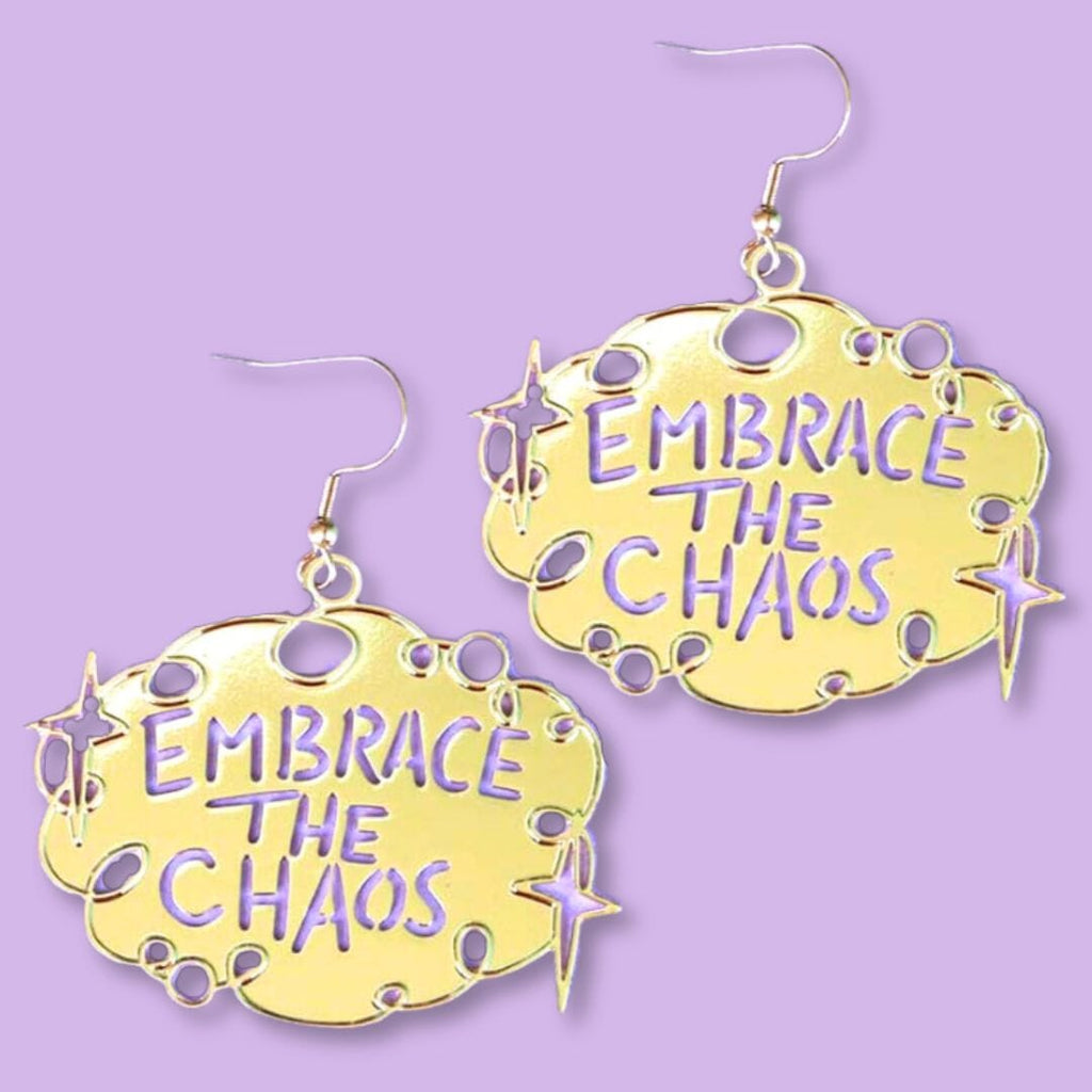 A pair of dangle brass earrings displayed on a purple background. The earrings read Embrace The Chaos.