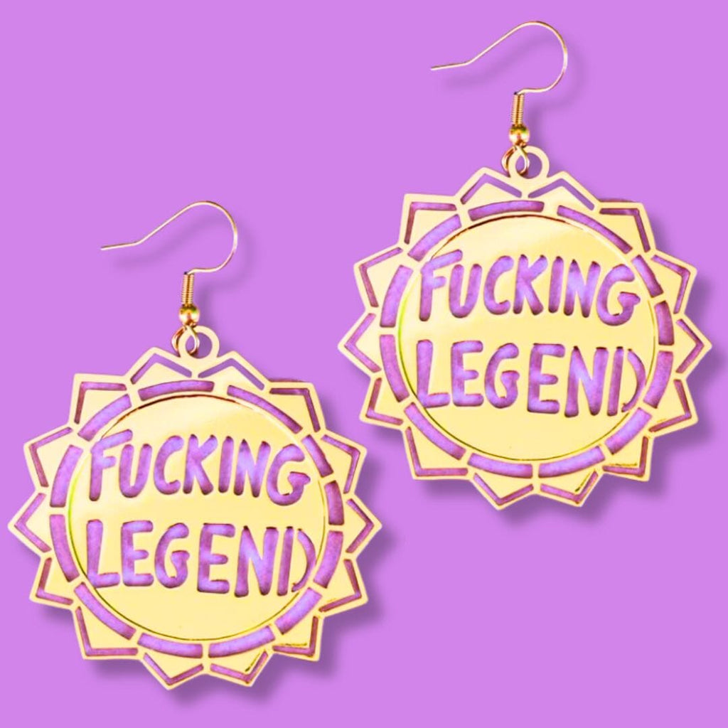 A pair of dangle brass earrings displayed on a purple background. The earrings read Fucking Legend.