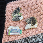Three pins on a knitted surface, including a corvid a cat, and a book with text.