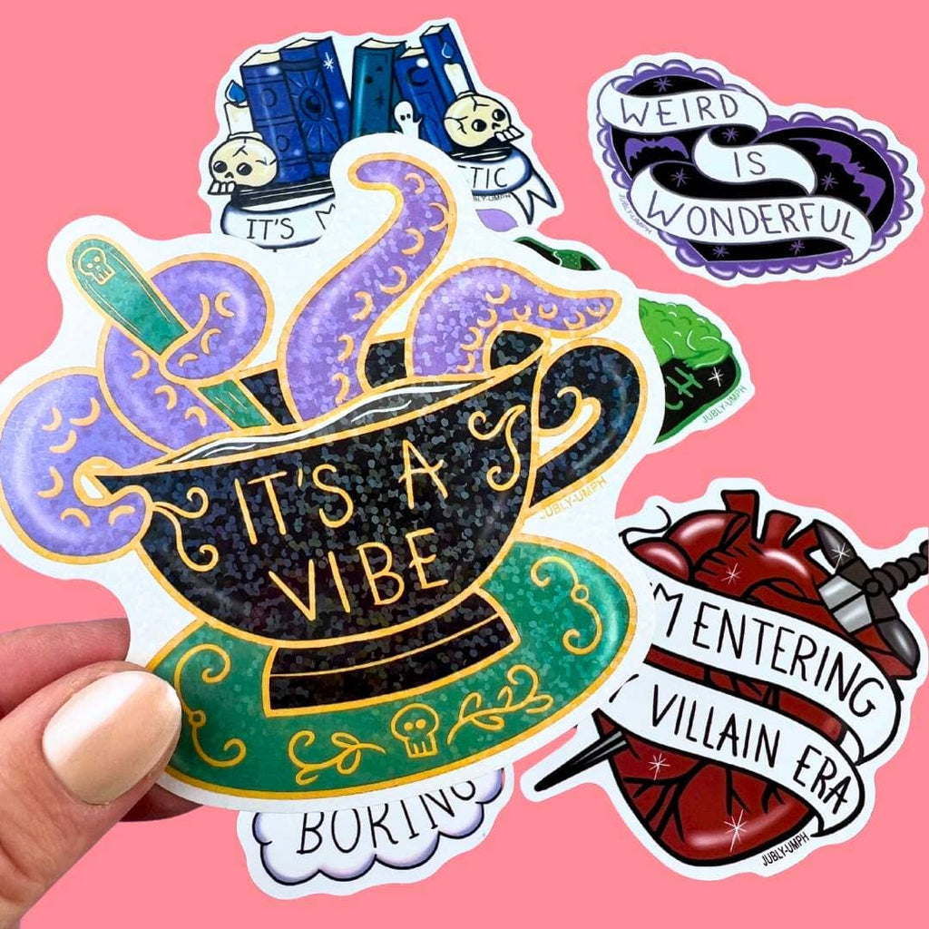 A vinyl sticker displayed in a hand. The sticker is in the shape of a black teacup with purple tenticles coming out. The pin reads It's A Vibe.