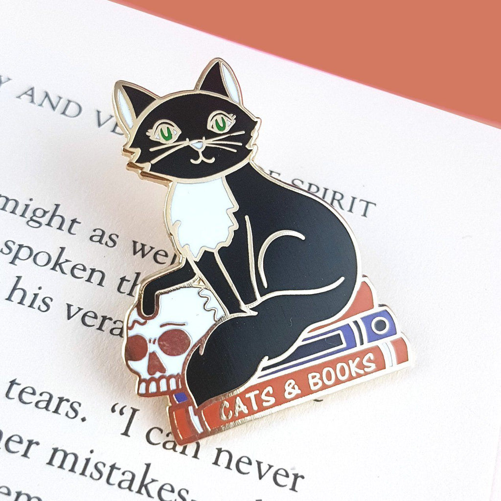 A hard enamel lapel pin. The pin is in the shape of a black and white cat with a scull, sitting on books. The pin reads Cat’s and Books.