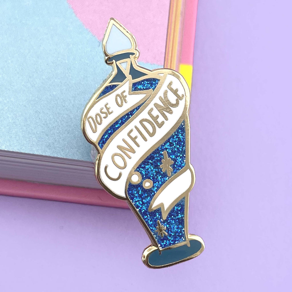 A hard enamel lapel pin on a purple background. The pin is in the profile of a bottle with dark blue glitter. There is a white ribbon around the bottle that reads Dose Of Confidence.