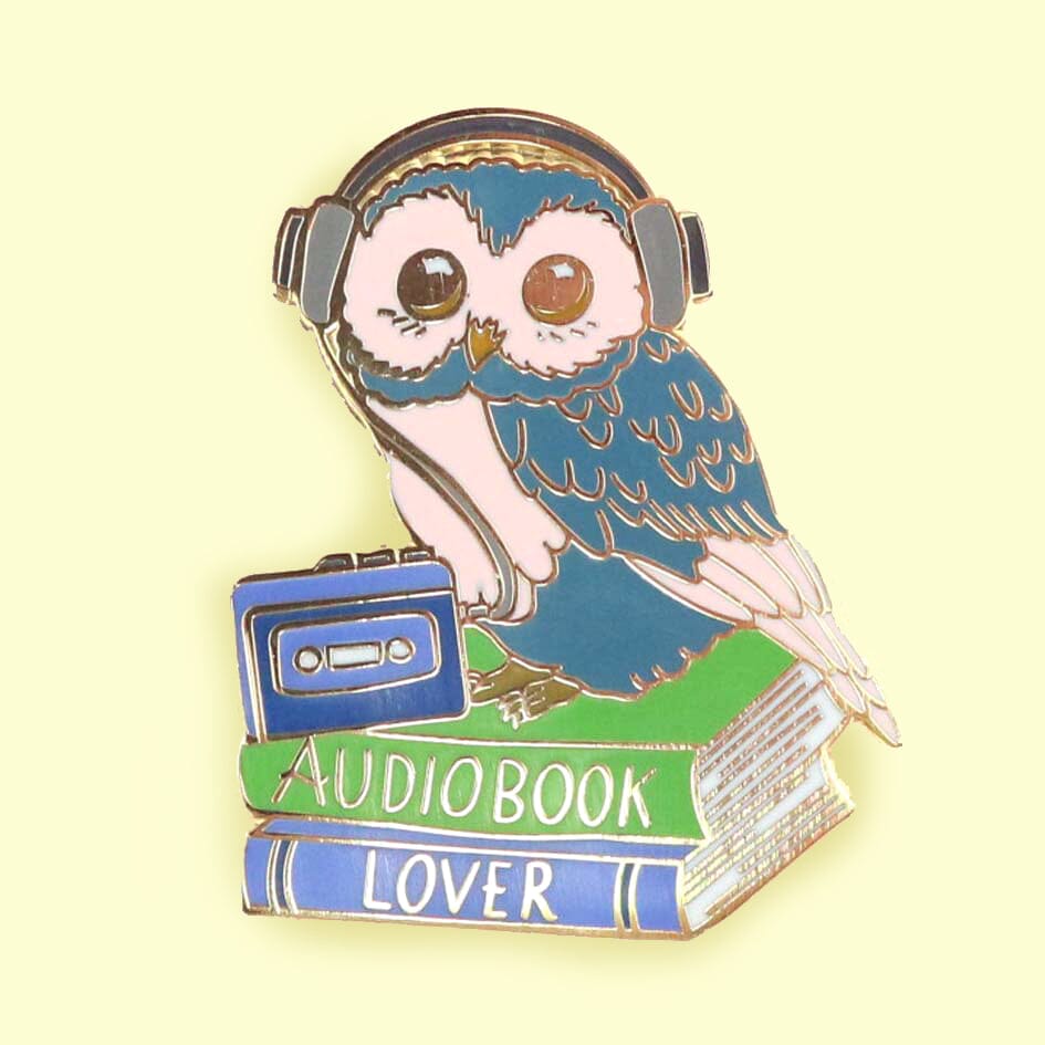A hard enamel lapel pin against a yellow background. The pin is in the shape of an owl sitting on a stack of books wearing headphones. The pin reads Audiobook Lover.
