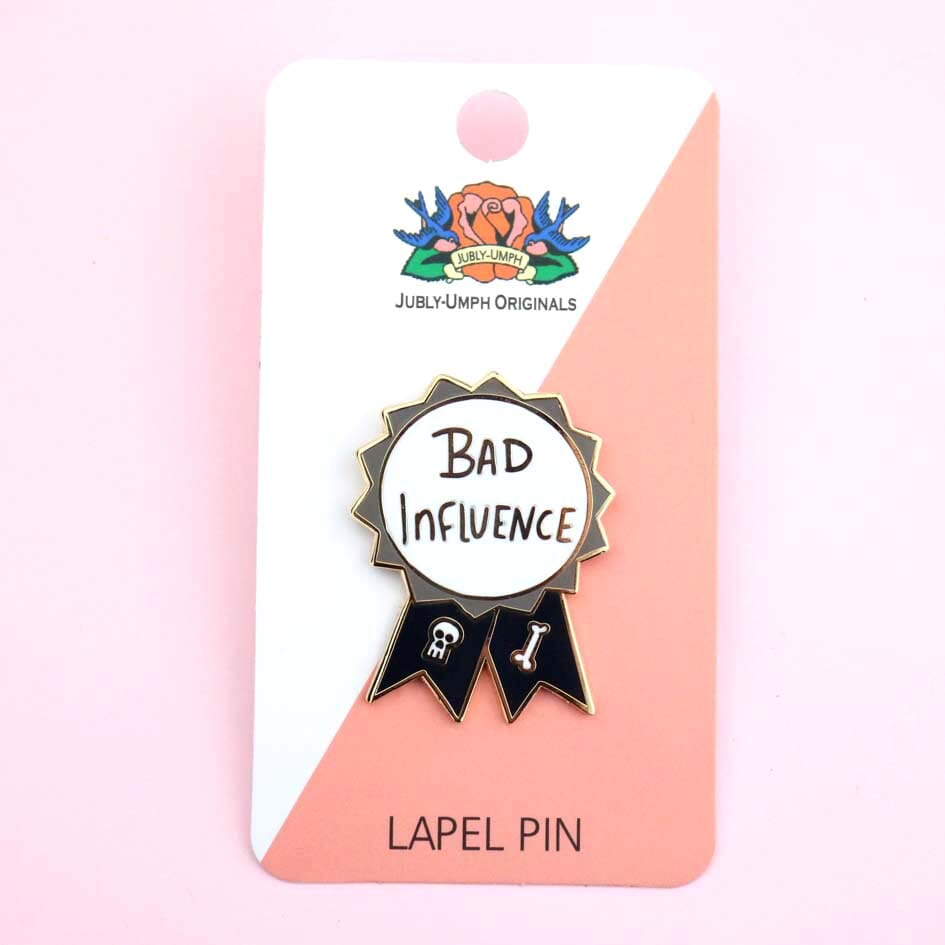 A hard enamel lapel pin on Jubly-Umph cardstock. The pin is black, white and gray in the shape of an award ribbon and reads Bad Influence.