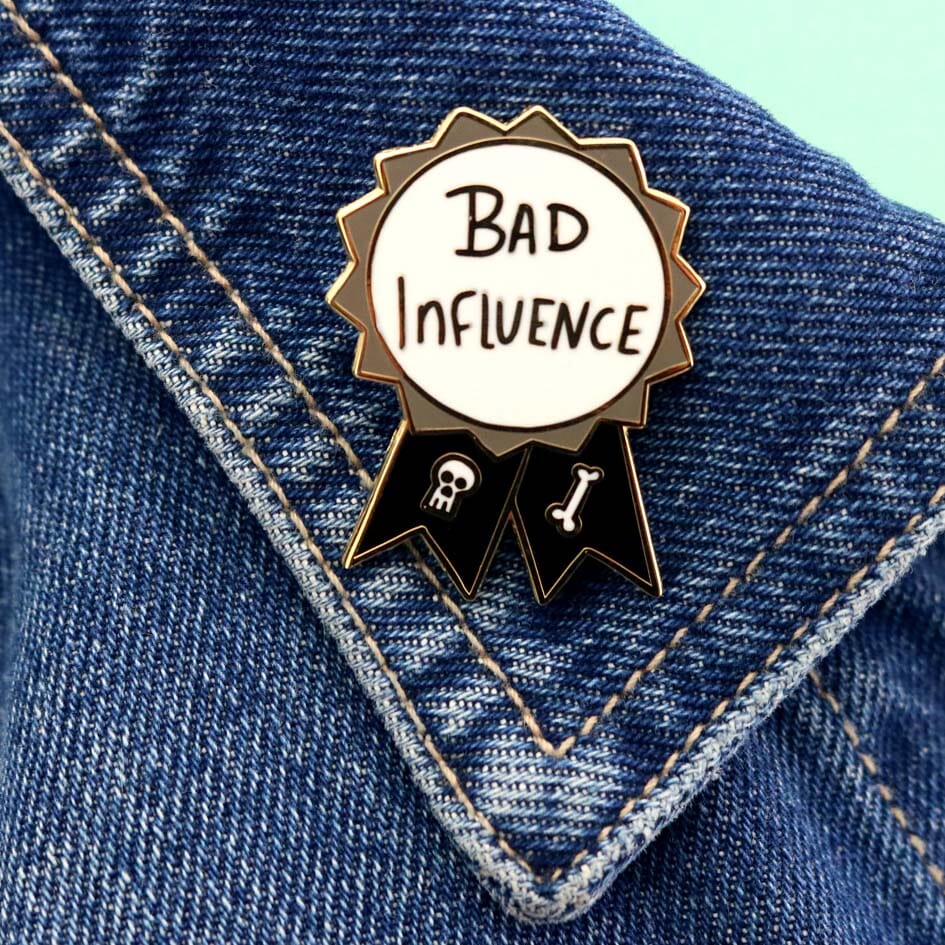 A hard enamel lapel pin against a denim jacket. The pin is black, white and gray in the shape of an award ribbon and reads Bad Influence.