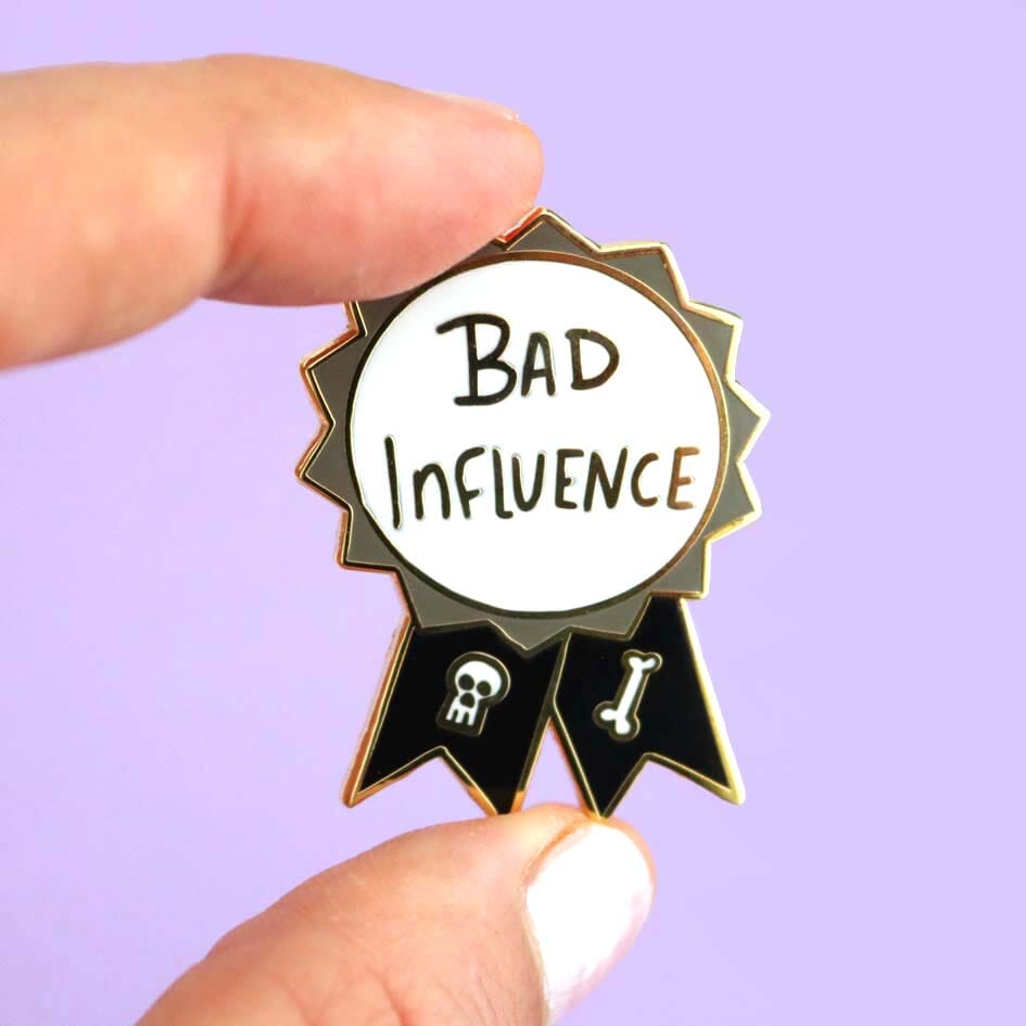 A hard enamel lapel pin being held in the hand against a purple background. The pin is black, white and gray in the shape of an award ribbon and reads Bad Influence.