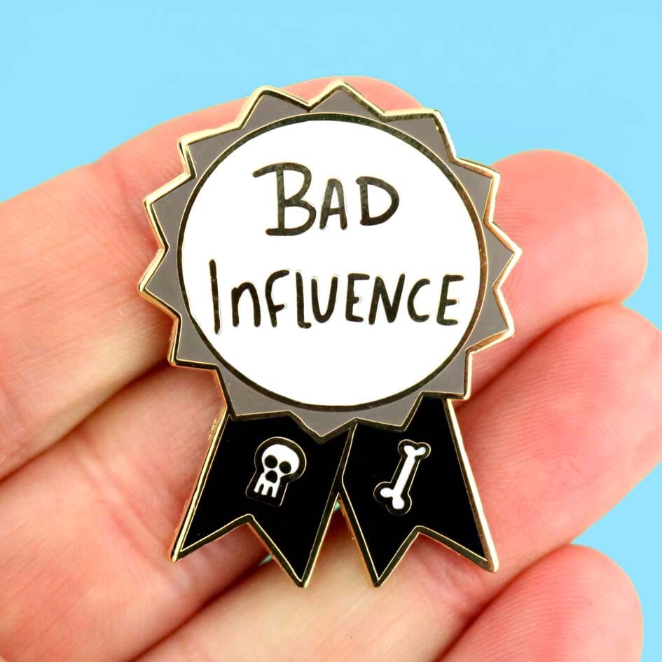 A hard enamel lapel pin being held in the hand against a blue background. The pin is black, white and gray in the shape of an award ribbon and reads Bad Influence.