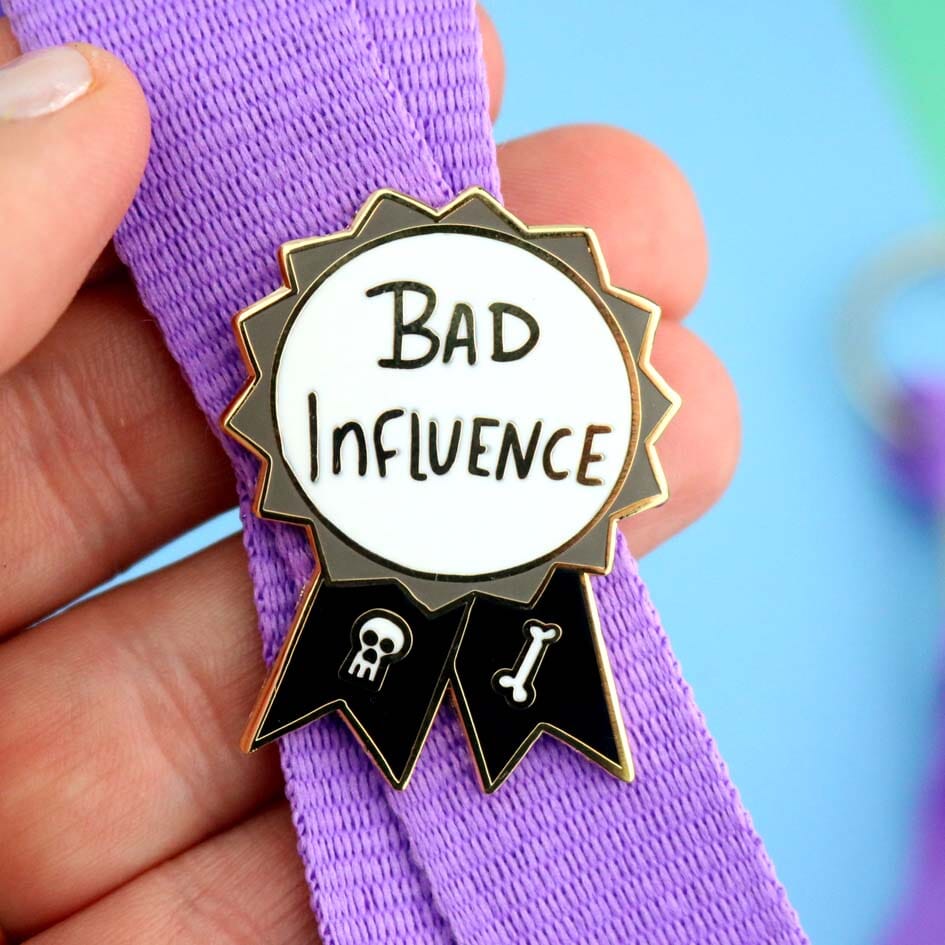 A hard enamel lapel pin being held in the hand on a lanyard. The pin is black, white and gray in the shape of an award ribbon and reads Bad Influence.