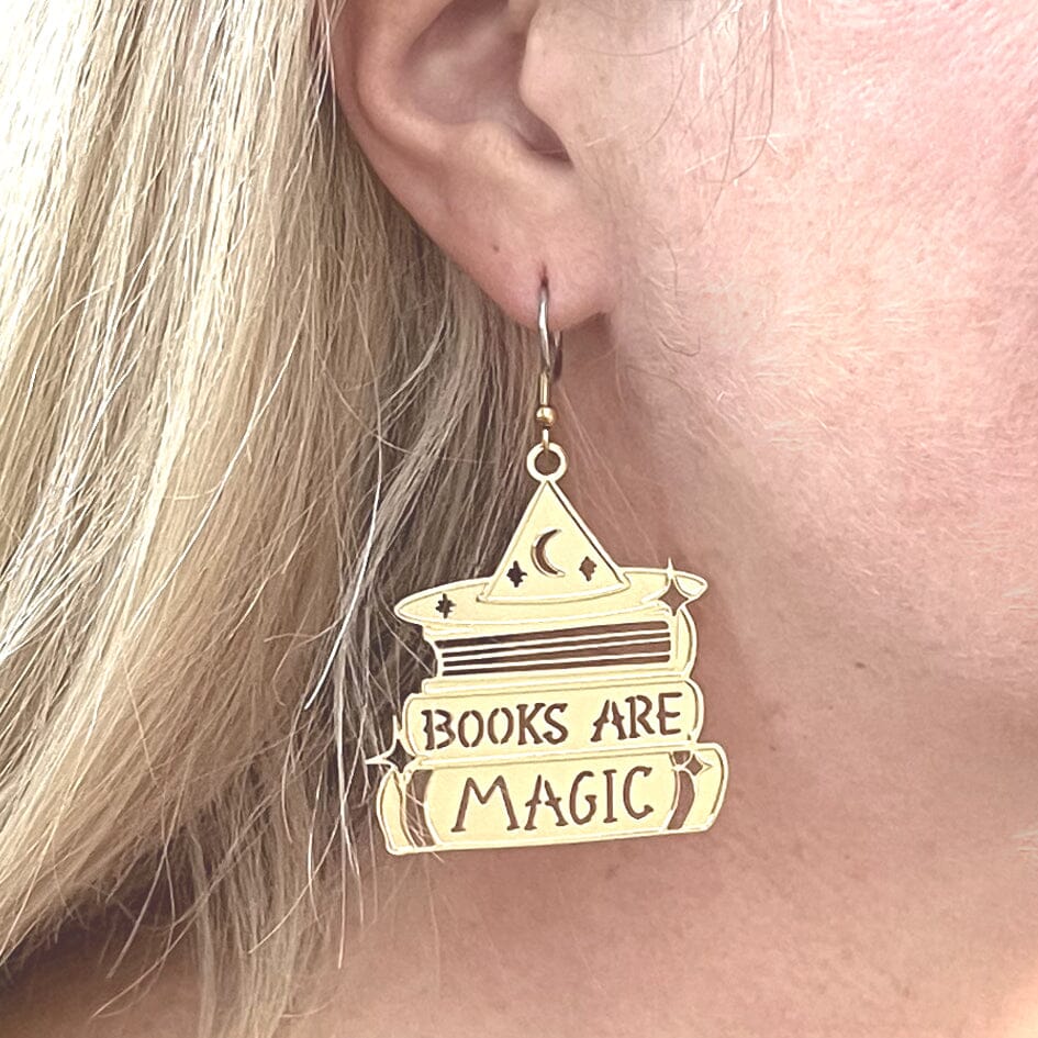 A single brass earring displayed from an ear lobe. The earring says Books are Magic with books and a witch’s hat. 