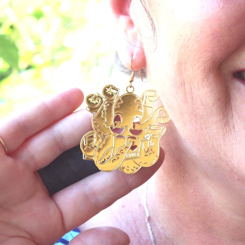 A single Brass earrings displayed from a woman’s ear. The earrings are of an octopus wearing glasses holding books in its tentacles.