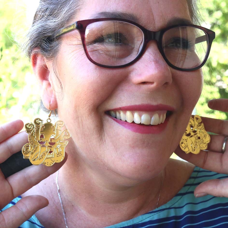 A pair of brass earrings displayed from a woman’s ears. The earrings are of an octopus wearing glasses holding books in its tentacles.