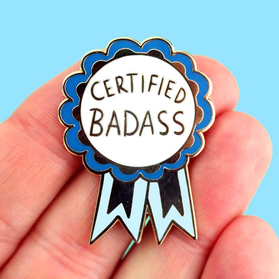 A hard enamel lapel pin being held in a hand. The pin is in the shape of a blue-ribbon award and reads Certified Badass.