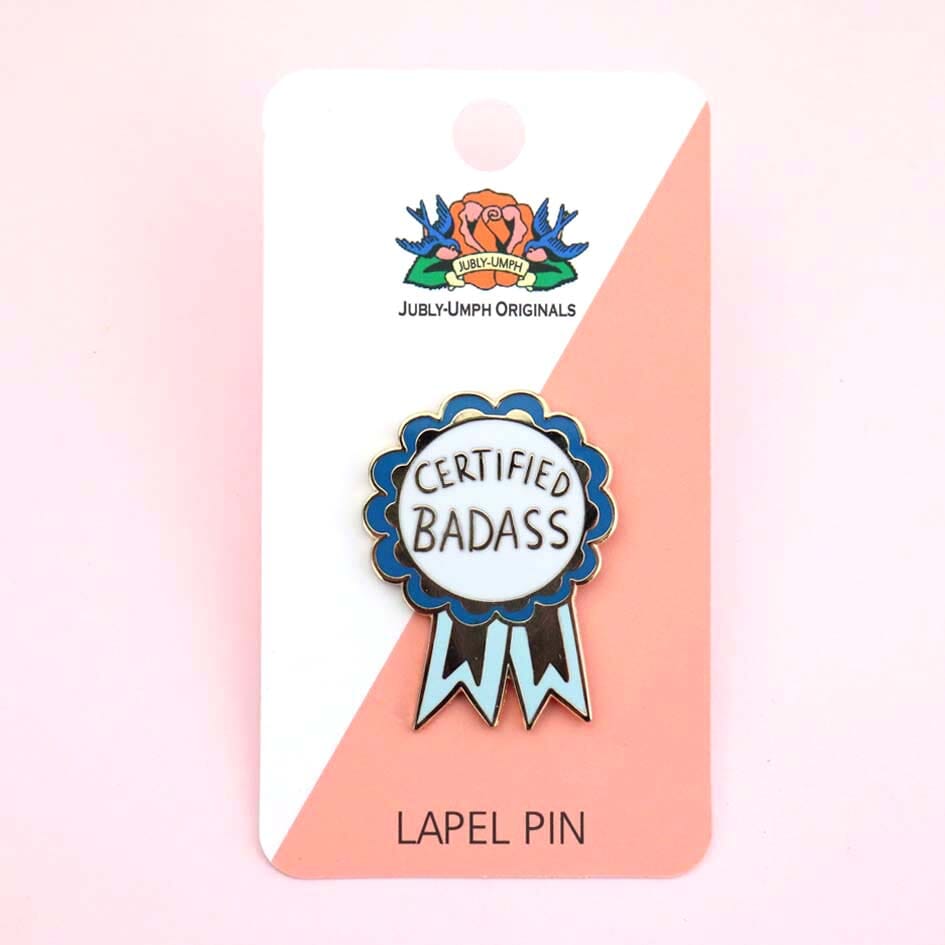 A hard enamel lapel pin on Jubly-Umph cardstock. The pin is in the shape of a blue-ribbon award and reads Certified Badass.