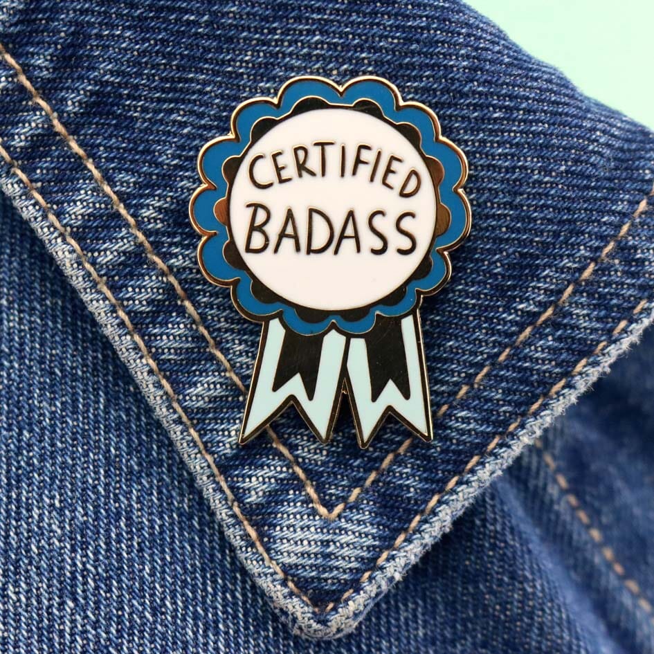 A hard enamel lapel pin on a denim jacket. The pin is in the shape of a blue-ribbon award and reads Certified Badass.