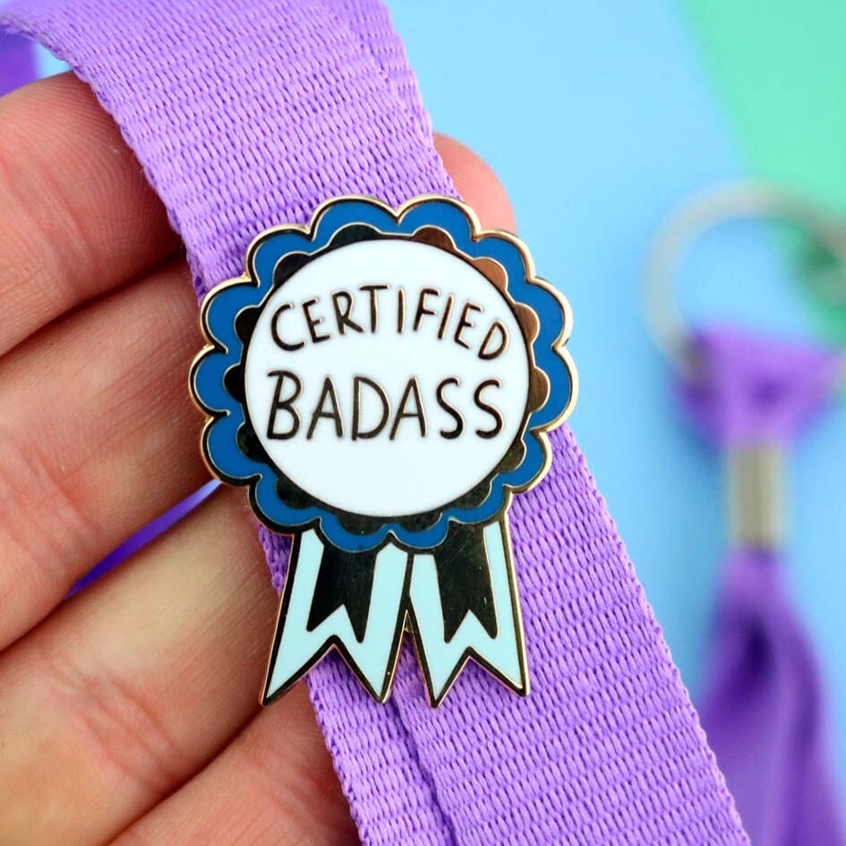 A hard enamel lapel pin on a lanyard. The pin is in the shape of a blue-ribbon award and reads Certified Badass.