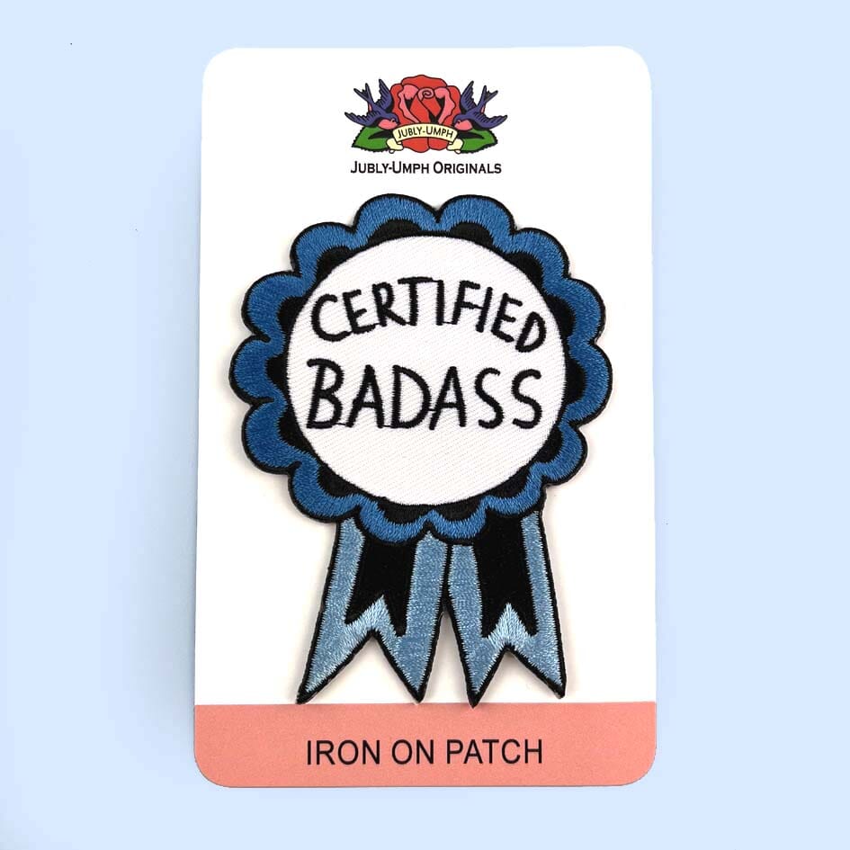 An iron on embroidered patch on Jubly-Umph cardstock against a blue background. The patch is in the shape of a blue-ribbon award and reads Certified Badass.