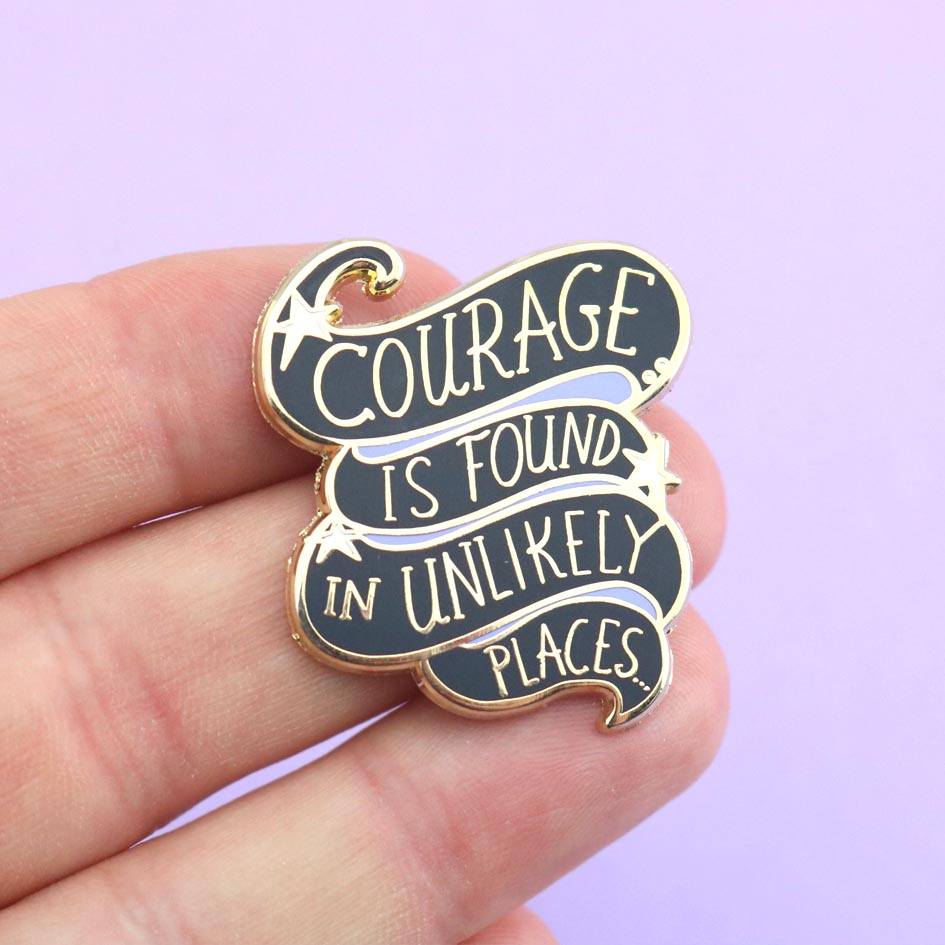 A hard enamel lapel pin being held in a hand. The pin is black and purple and reads Courage Is Found In Unlikely Places.