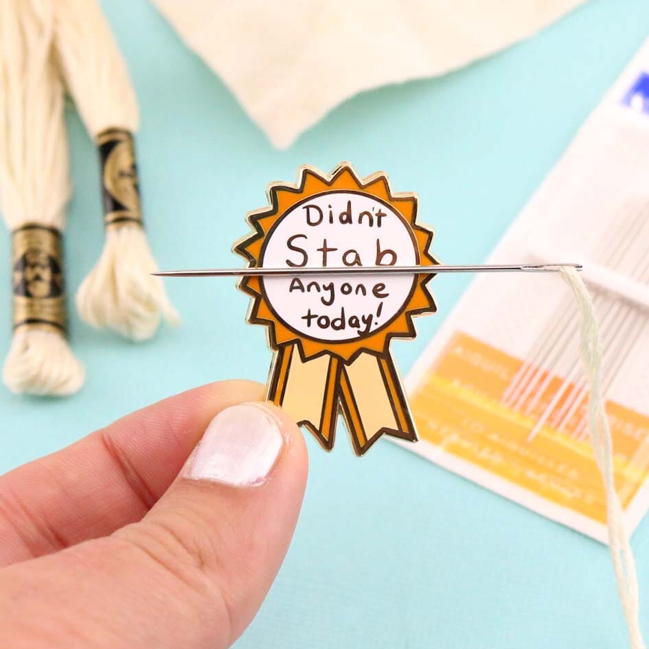 A hard enamel magnetic needle minder being held in a hand with a suspended needle and thread. The pin is in the shape of an award ribbon. The ribbon is yellow and white, and reads Didn’t Stab Anyone Today!