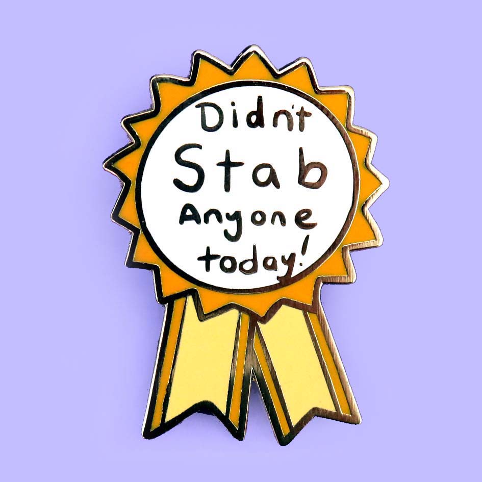 A hard enamel lapel pin on a purple background. The pin is in the shape of an award ribbon. The ribbon is yellow and white, and reads Didn’t Stab Anyone Today!