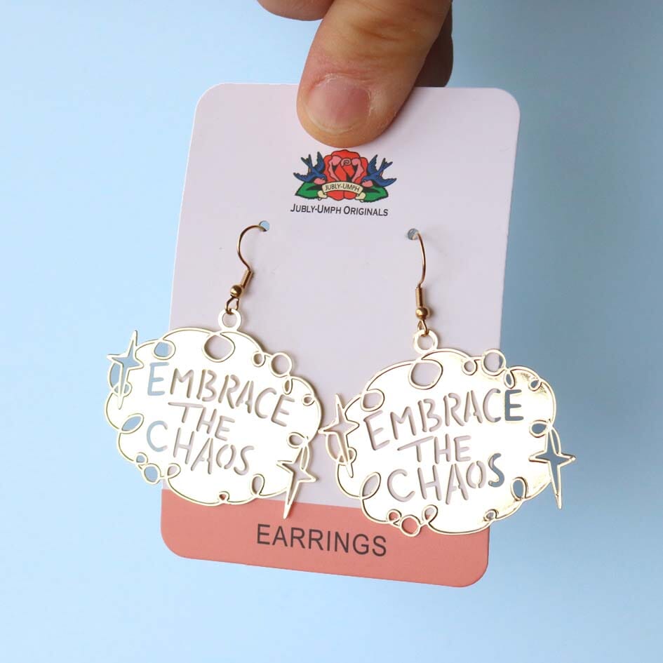 A pair of dangle brass earrings displayed on Jubly-Umph cardstock. The earrings read Embrace The Chaos.