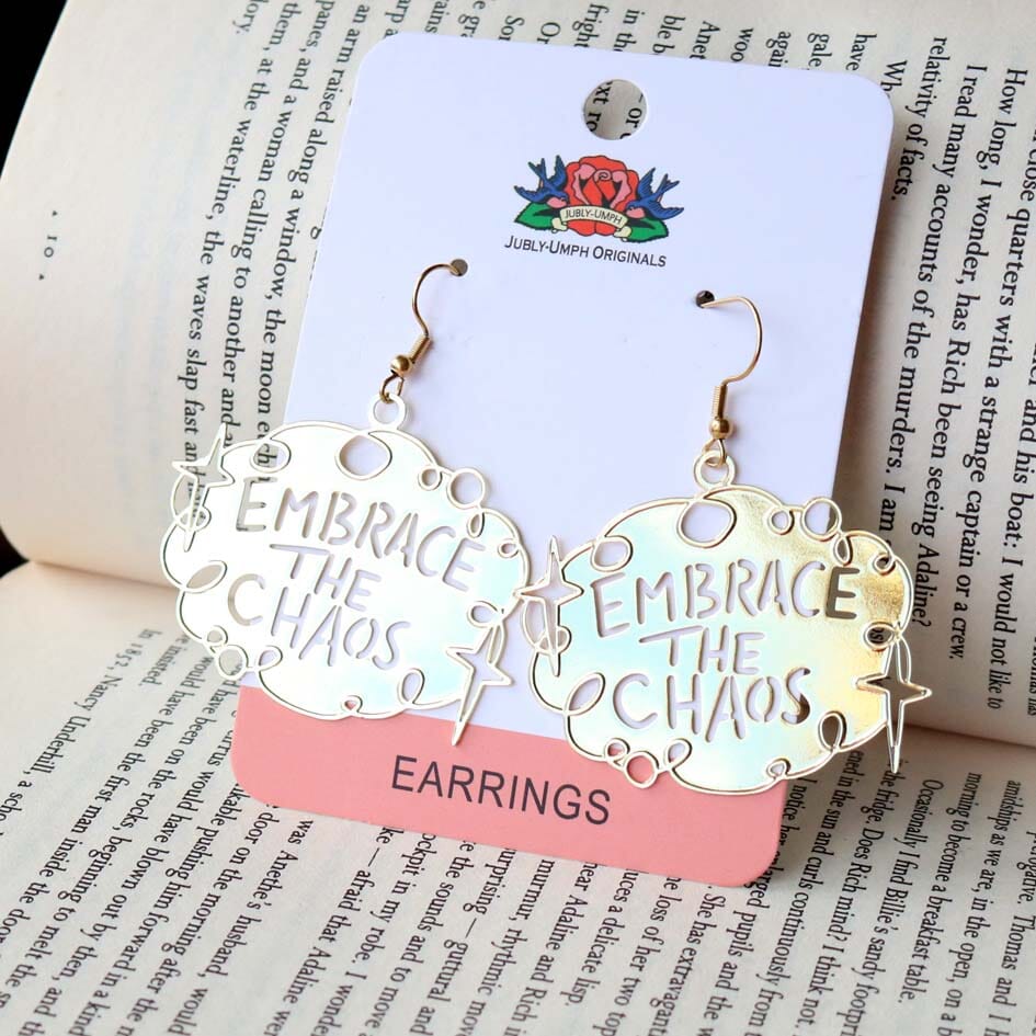 A pair of dangle brass earrings displayed on Jubly-Umph cardstock. The earrings read Embrace The Chaos.