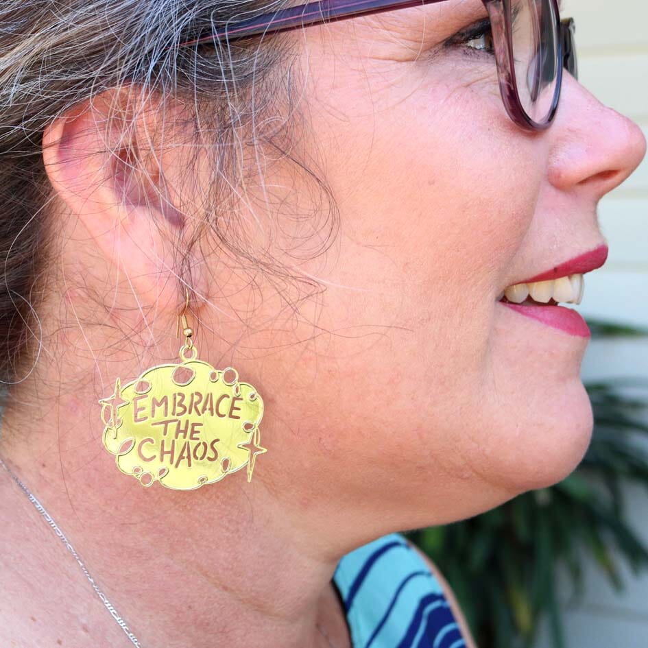 A dangle brass earring being worn by a smiling woman. The earrings reads Embrace The Chaos.