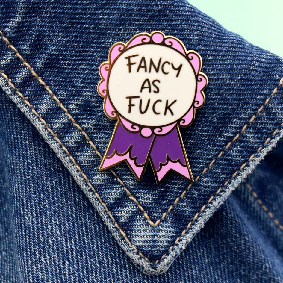 A hard enamel lapel pin on a denim jacket. The pin is in the shape of a pink and purple ribbon award and reads Fancy As Fuck.