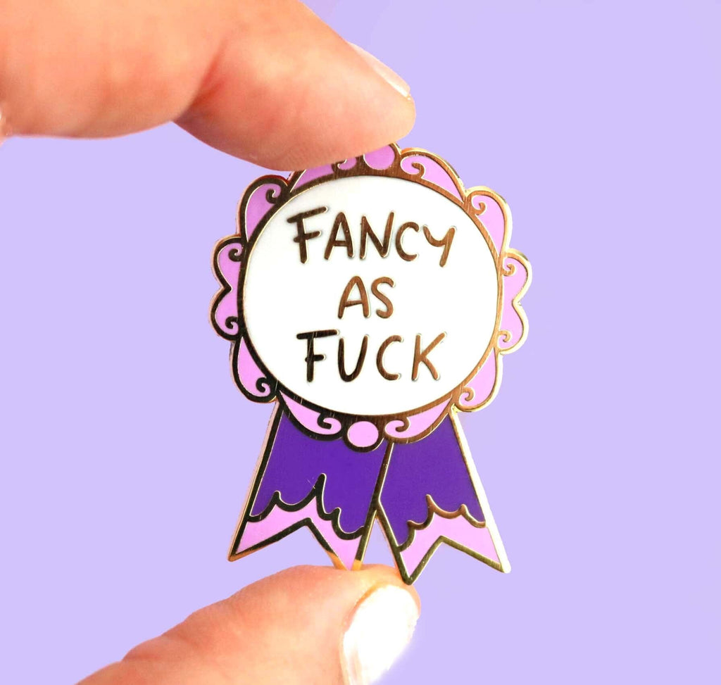 A hard enamel lapel pin being held in a hand. The pin is in the shape of a pink and purple ribbon award and reads Fancy As Fuck.