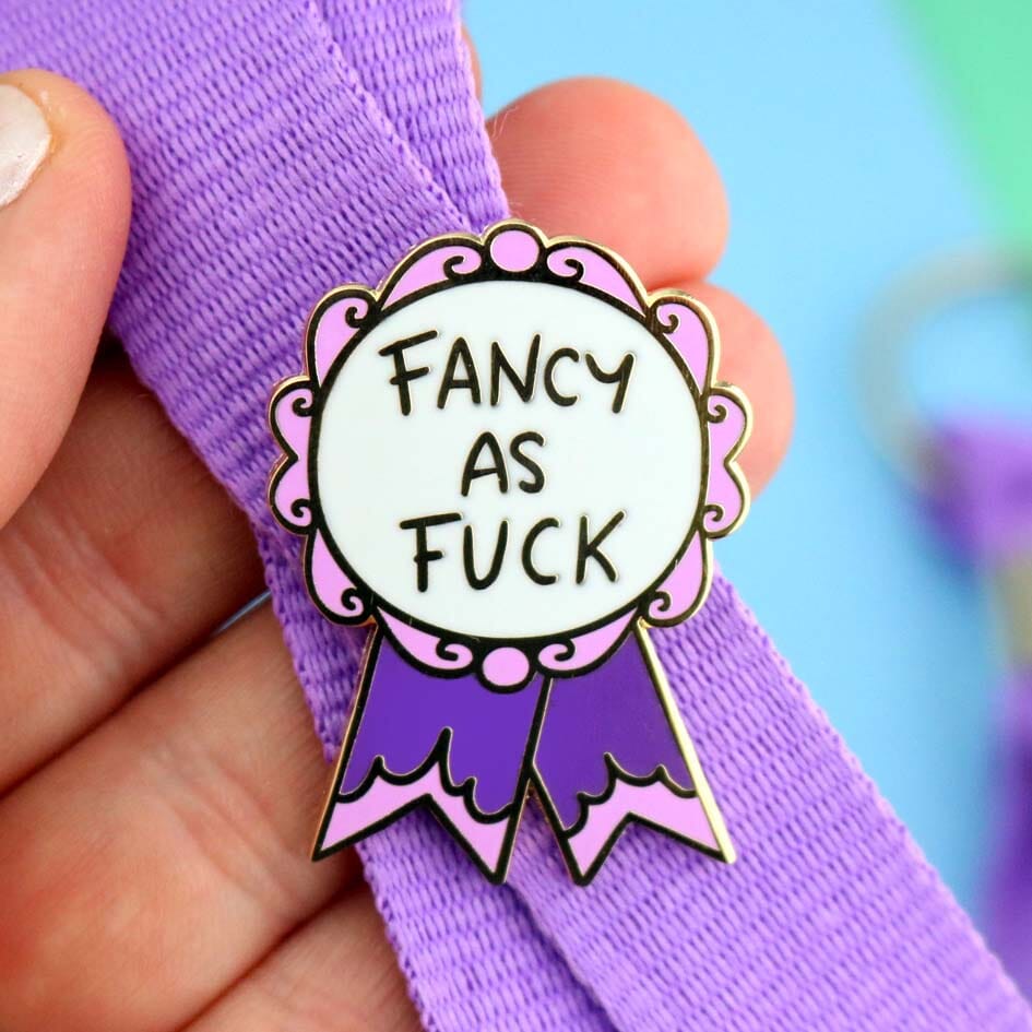 A hard enamel lapel pin on a purple lanyard. The pin is in the shape of a pink and purple ribbon award and reads Fancy As Fuck.