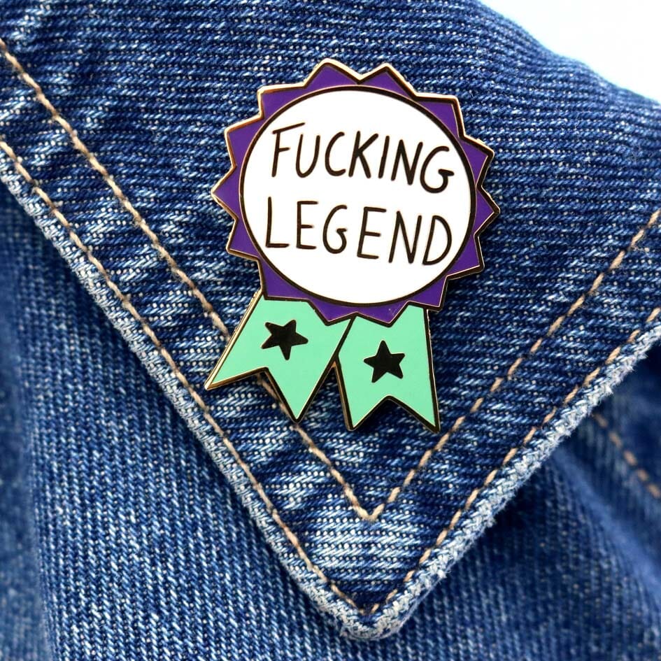 A hard enamel lapel pin being displayed on a denim jacket. The pin is in the shape of a teal and purple ribbon award and reads Fucking Legend.