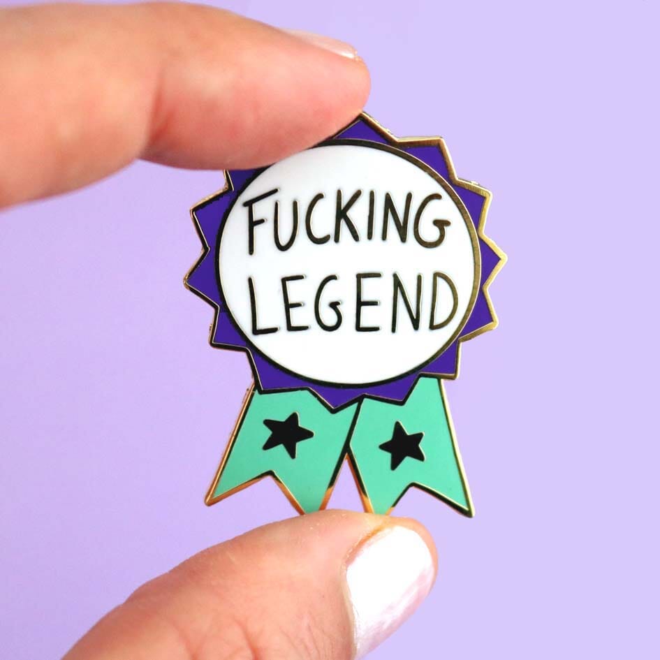A hard enamel lapel pin being held in a hand. The pin is in the shape of a teal and purple ribbon award and reads Fucking Legend