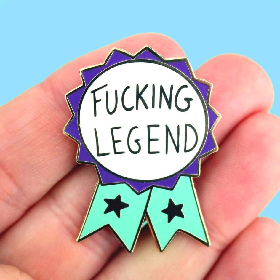 A hard enamel lapel pin being held in a hand. The pin is in the shape of a teal and purple ribbon award and reads Fucking Legend.