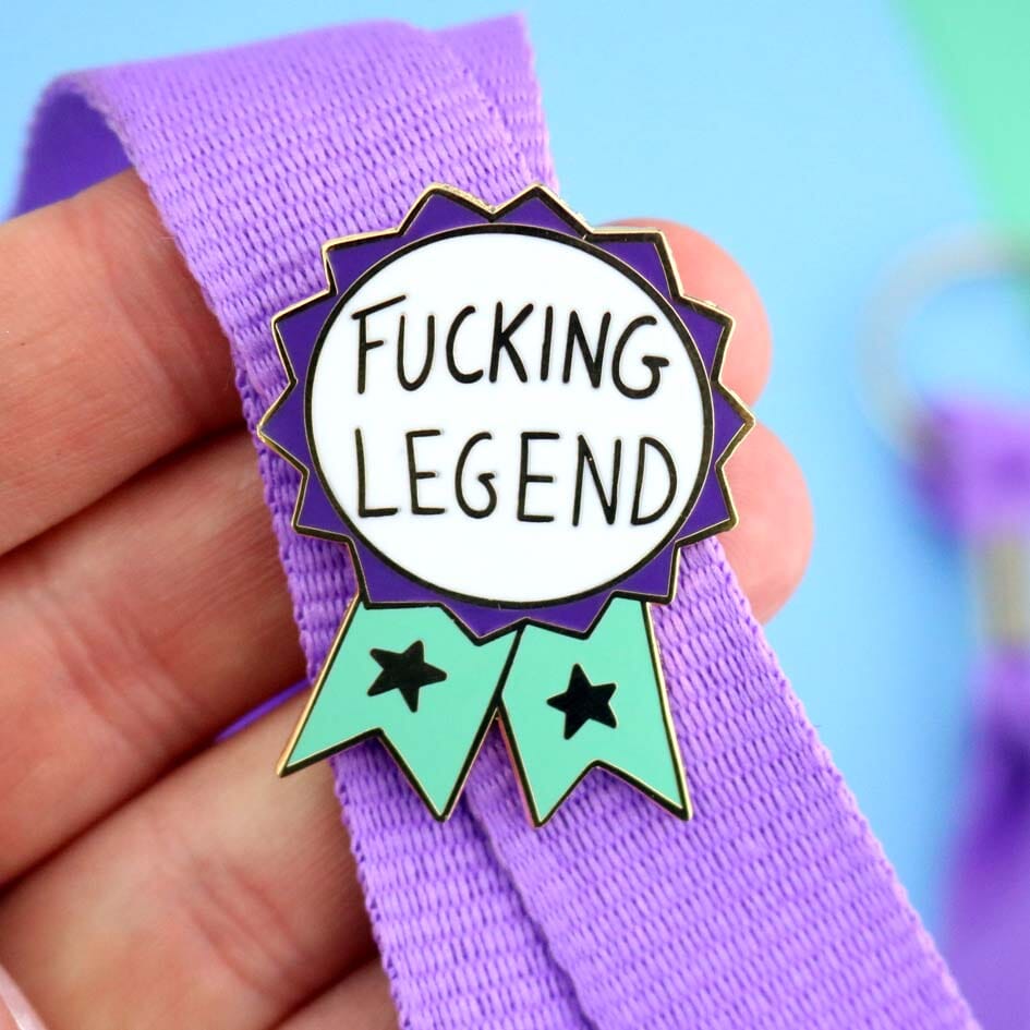A hard enamel lapel pin on a purple lanyard. The pin is in the shape of a teal and purple ribbon award and reads Fucking Legend.