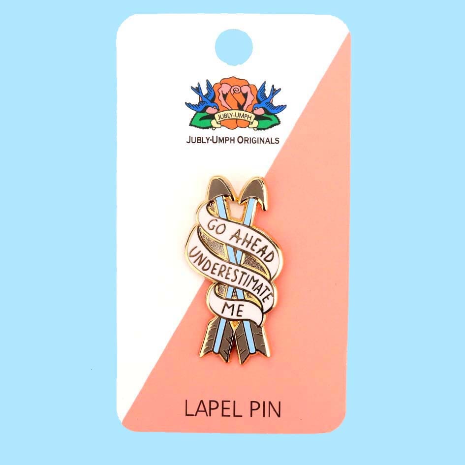 A hard enamel lapel pin on Jubly-Umph backing card. The pin is in the shape of two arrows and reads Go Ahead Underestimate Me.