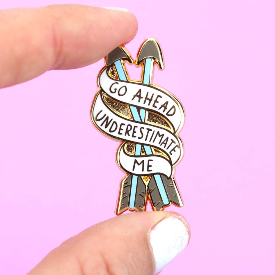A hard enamel lapel pin being held in a hand. The pin is in the shape of two arrows and reads Go Ahead Underestimate Me.