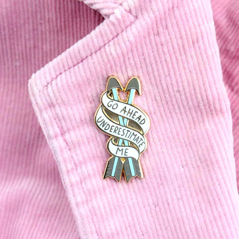 A hard enamel lapel pin on a pink jacket. The pin is in the shape of two arrows and reads Go Ahead Underestimate Me.