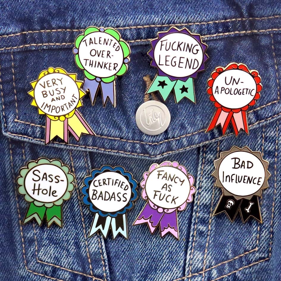 Eight hard enamel pins are attached to a lanyard. They are each in the shape of an award ribbon. They read Sass-Hole, Very Busy and Important, Bad Influence, Fancy As Fuck, Fucking Legend, Un-apologetic, Talented Overthinker, Certified Badass.