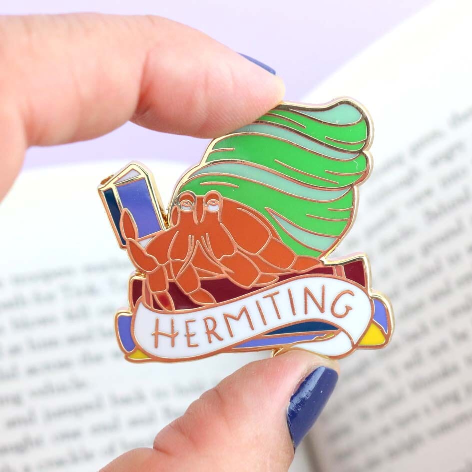 A hard enamel lapel pin being held in a hand. The pin is in the shape of a Hermit Crab reading a book. The pin says Hermit Crab.