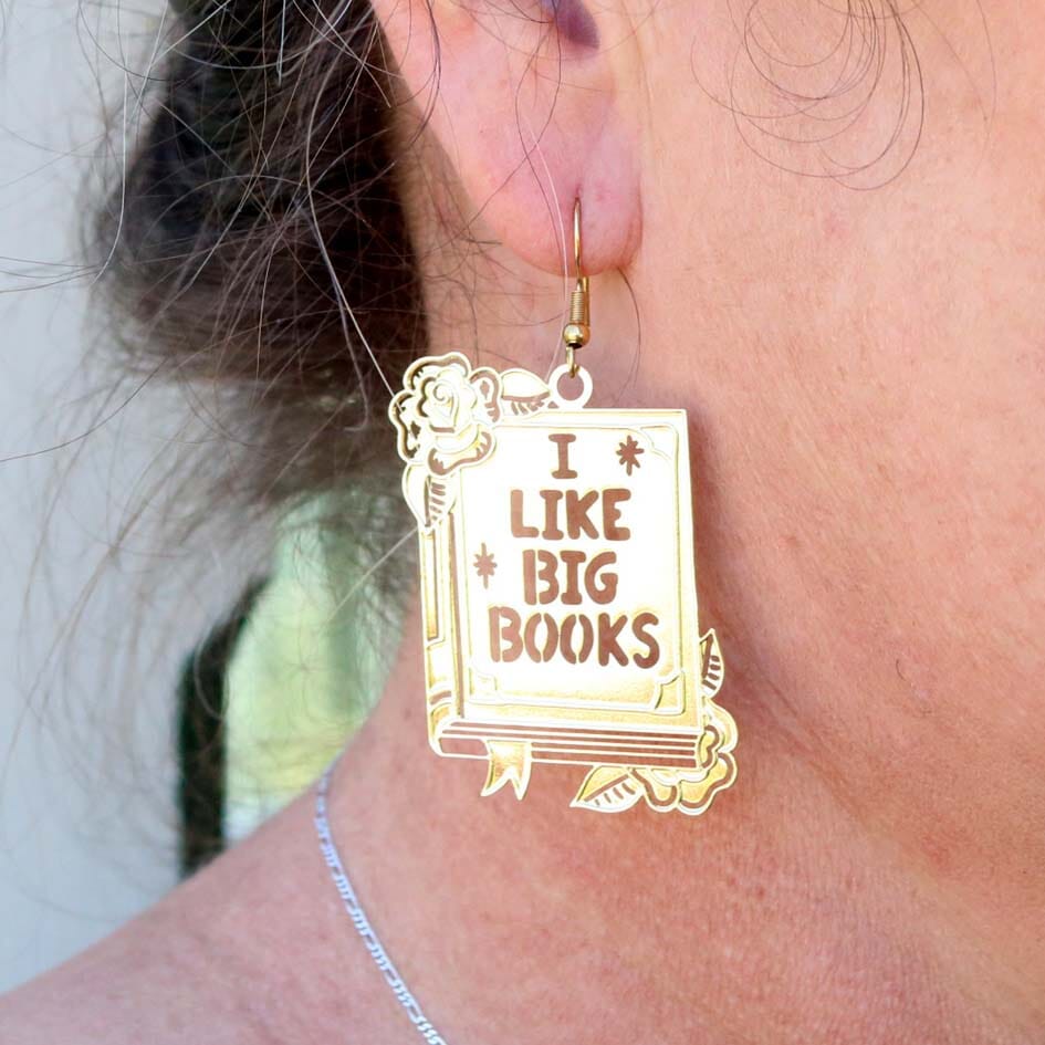 A single brass earring being worn by a model. The earring reads I Like Big Books.