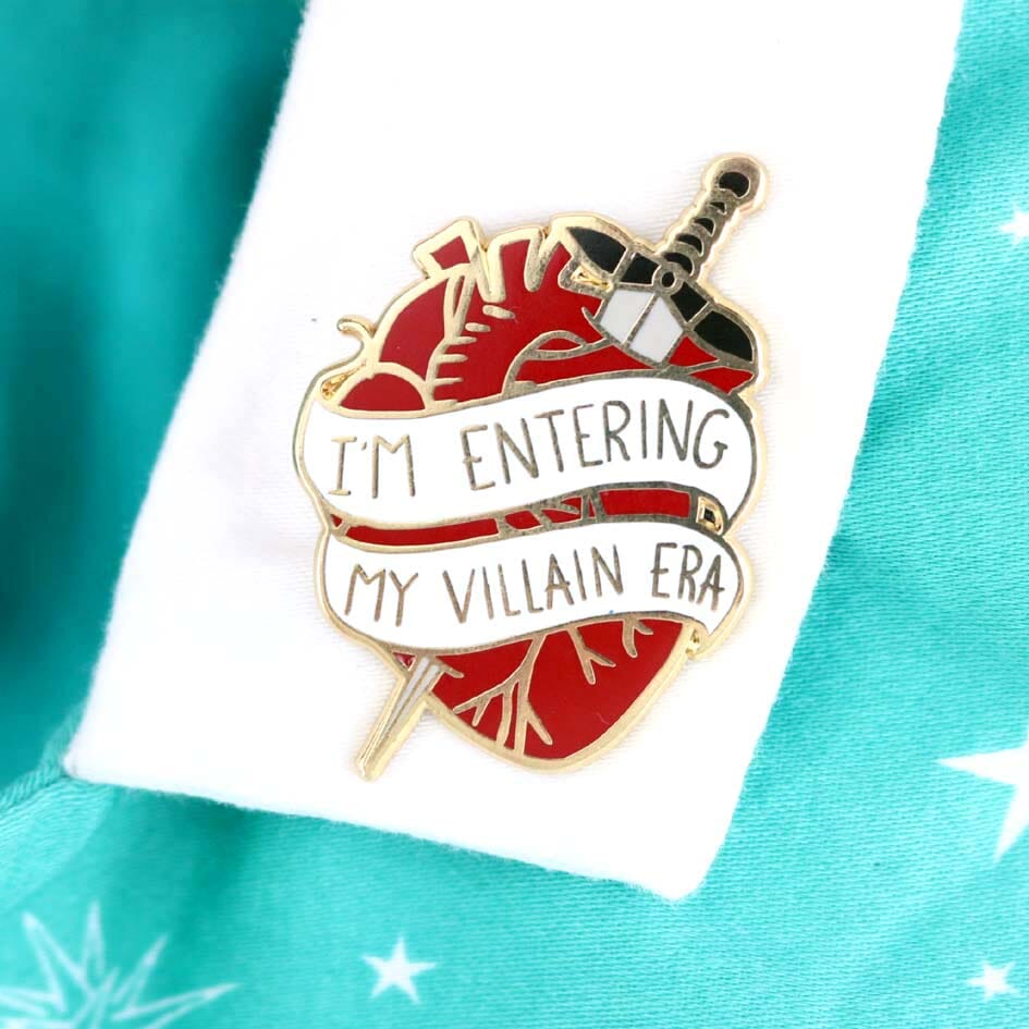 A hard enamel lapel pin being modelled on a white collar. The lapel pin is in the shape of a human heart with a dagger. The pin reads I'm Entering My Villain Era.