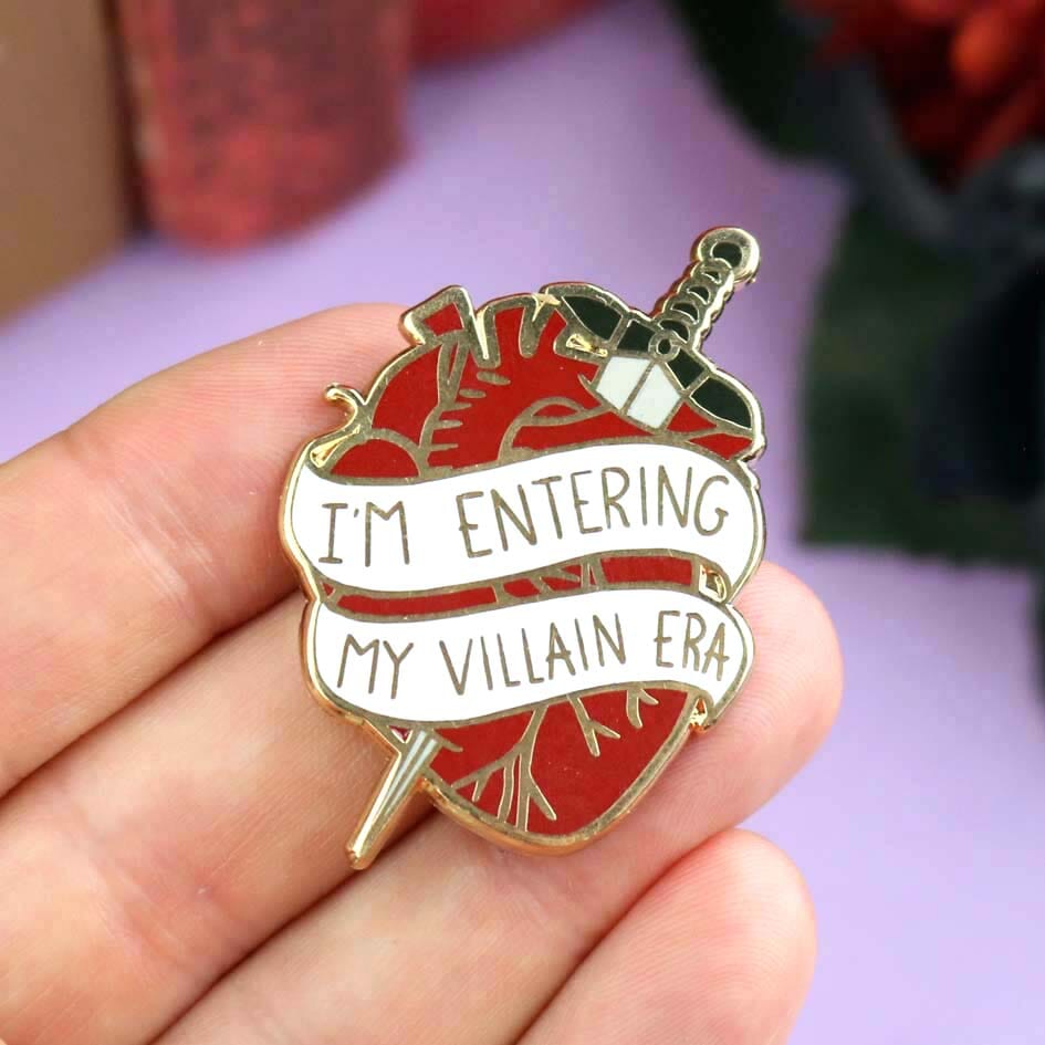 A hard enamel lapel pin being held in a hand. The lapel pin is in the shape of a human heart with a dagger. The pin reads I'm Entering My Villain Era.