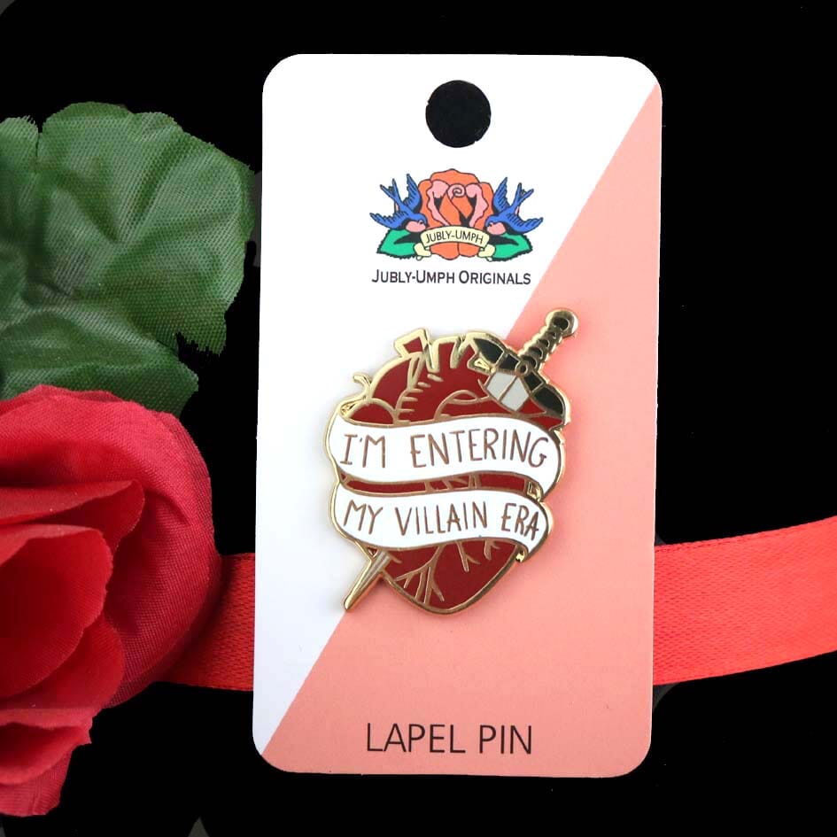 A hard enamel lapel pin displayed on a Jubly-Umph backing card. The lapel pin is in the shape of a human heart with a dagger. The pin reads I'm Entering My Villain Era.
