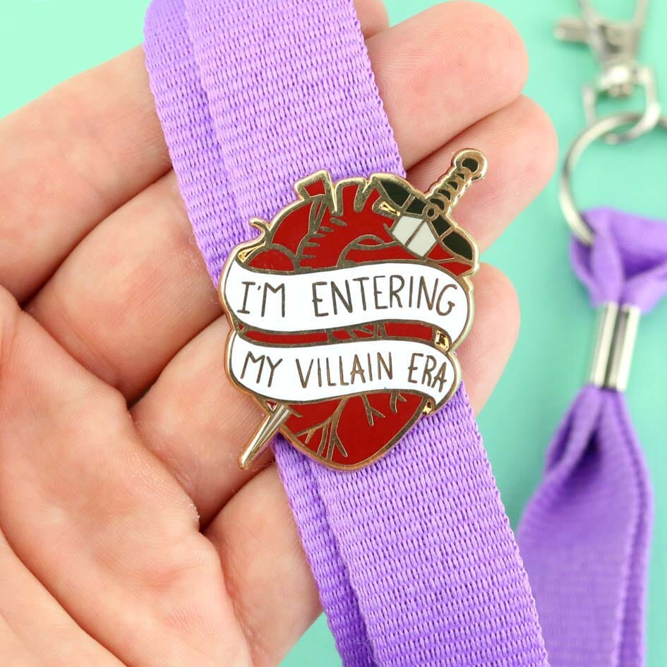 A hard enamel lapel displayed on a purple lanyard. The lapel pin is in the shape of a human heart with a dagger. The pin reads I'm Entering My Villain Era.