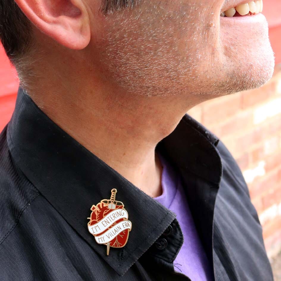 A hard enamel lapel pin being modelled on a black shirt. The lapel pin is in the shape of a human heart with a dagger. The pin reads I'm Entering My Villain Era.