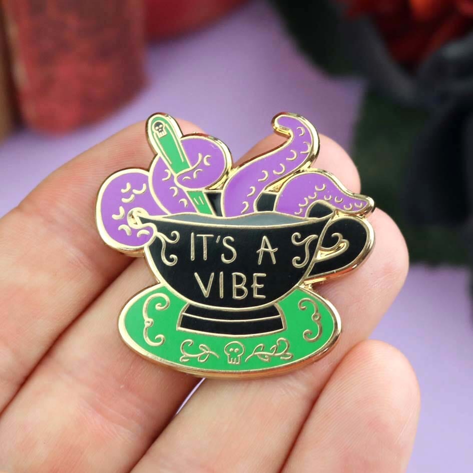 A hard enamel lapel pin being held in a hand. The lapel pin is in the shape of a black teacup with purple tenticles coming out. The pin reads It's A Vibe.