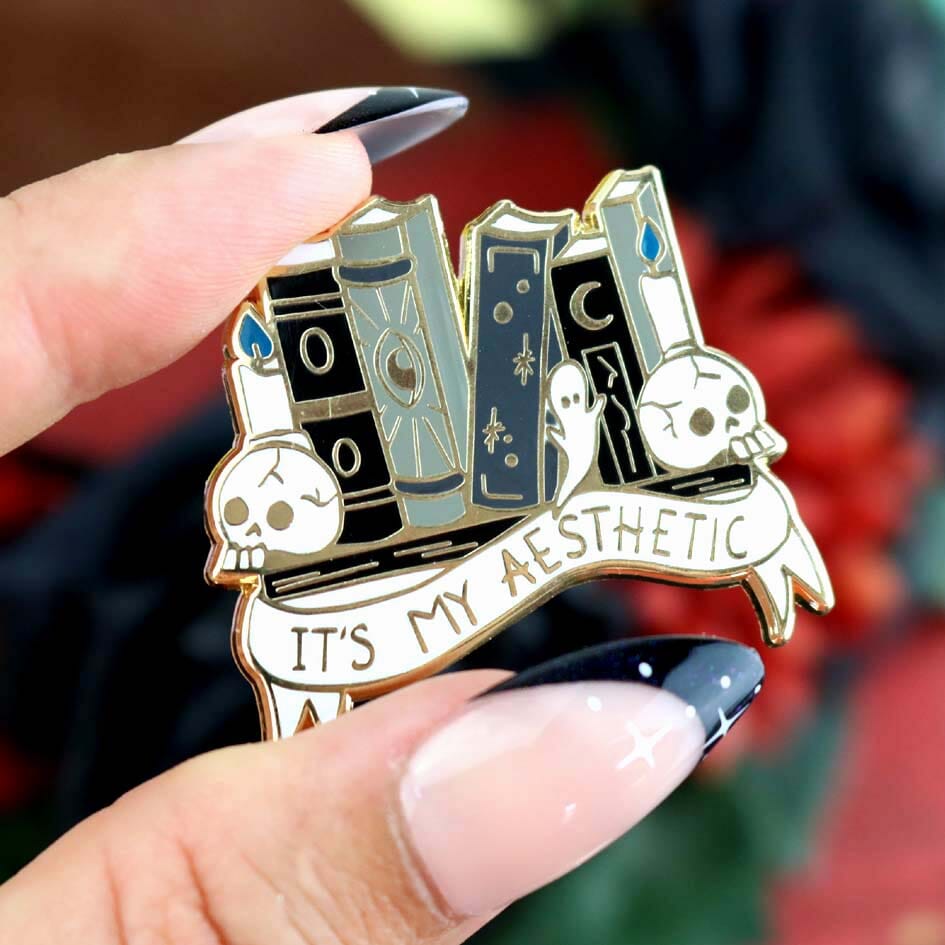 A hard enamel lapel pin being held in a hand. The lapel pin is in the shape of books on a shelf with skulls and candles. The pin reads It's My Aesthetic.